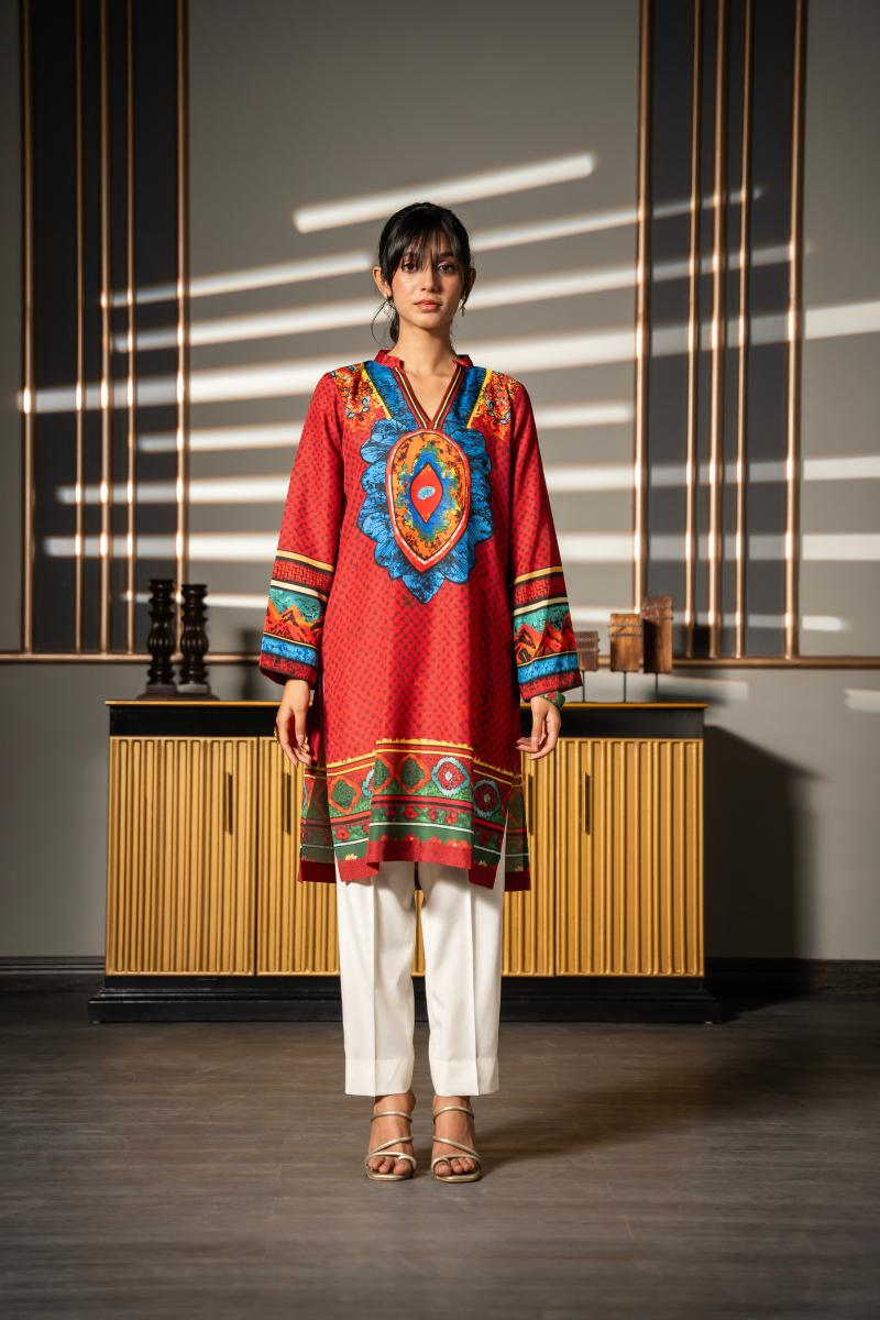 A woman stands in a modern interior wearing a red embroidered tunic with colorful geometric patterns, white pants, and beige sandals. The background features a wooden cabinet and wall panels with dramatic window light shadows.