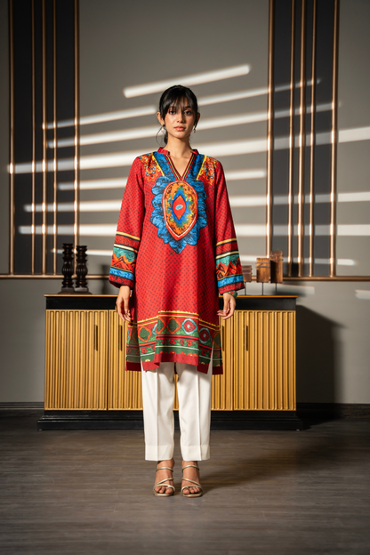 A woman stands in a modern interior wearing a red embroidered tunic with colorful geometric patterns, white pants, and beige sandals. The background features a wooden cabinet and wall panels with dramatic window light shadows.