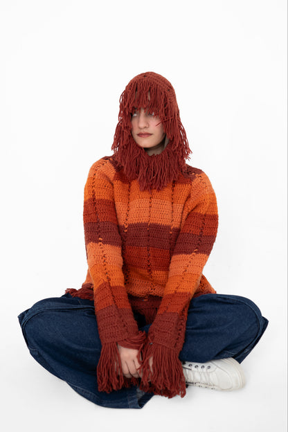 Model seated cross-legged wearing the Afterglow crochet hoodie with its long fringed hood pulled over her face