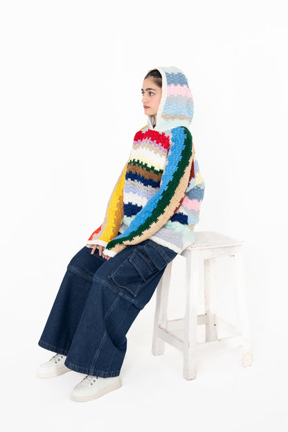 Model seated on a white stool wearing the Colour Out Loud multicolour crochet hoodie with the hood up
