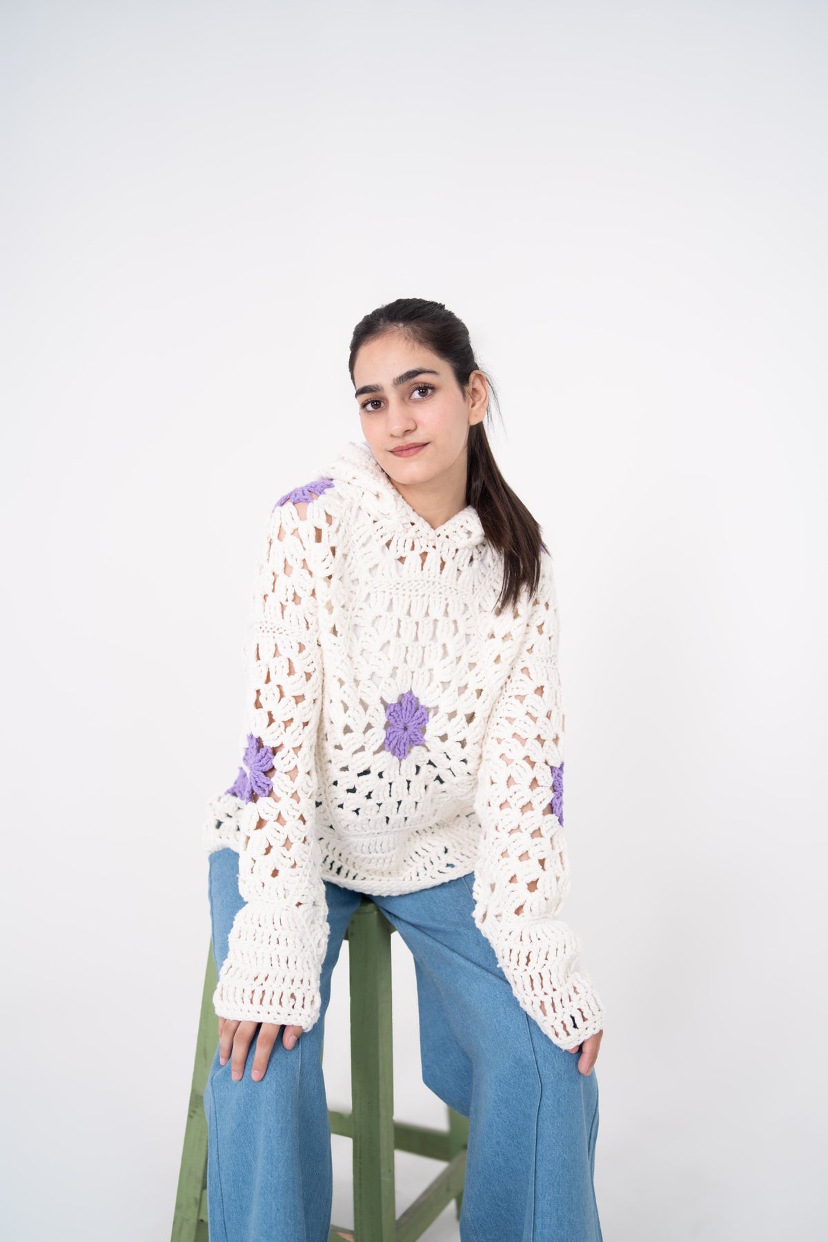 Lilac Cameo handmade ivory openwork crochet hoodie with lavender floral motifs, model seated on a green stool